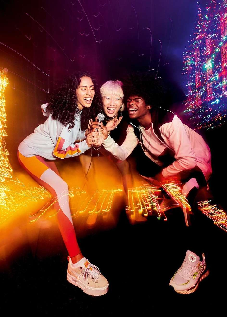 Three young adults joyfully sing into a shared microphone against a blurred backdrop, styled in comfortable athletic wear, capturing a lively moment.