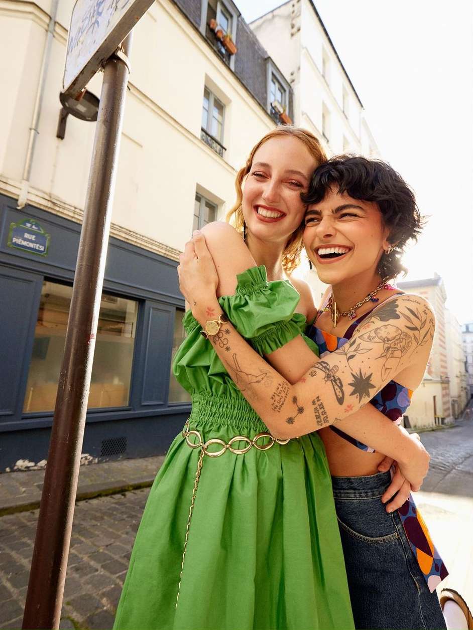 Two adult women smiling and embracing on a city street, one in a green dress and the other in a patterned top and jeans.