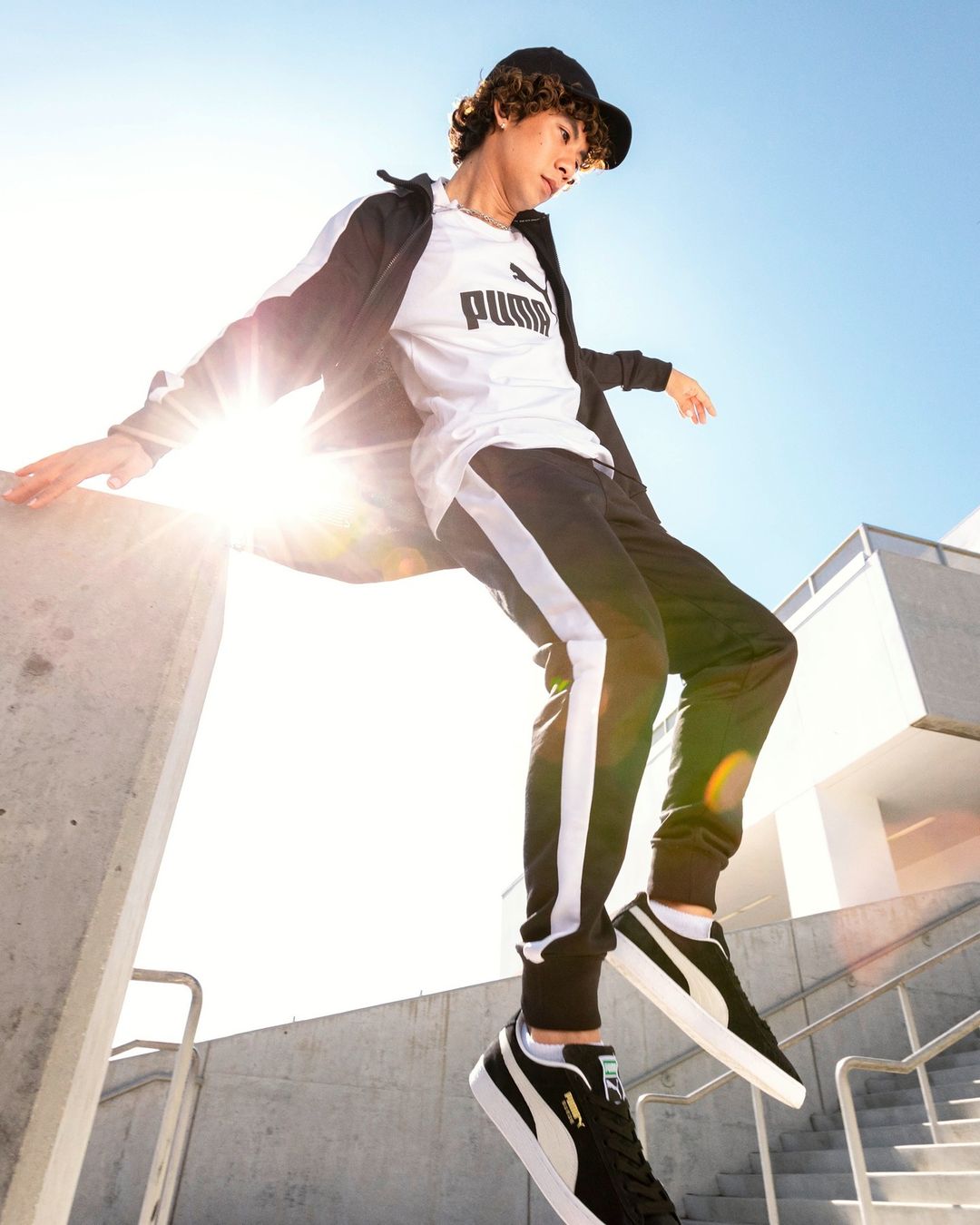 Young adult poses on a concrete ledge in Puma branded activewear and sneakers against a blue sky with a building in the background.