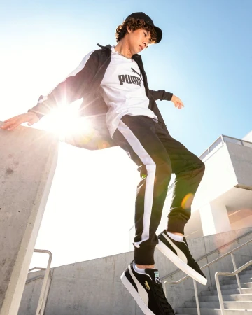 Young adult poses on a concrete ledge in Puma branded activewear and sneakers against a blue sky with a building in the background.