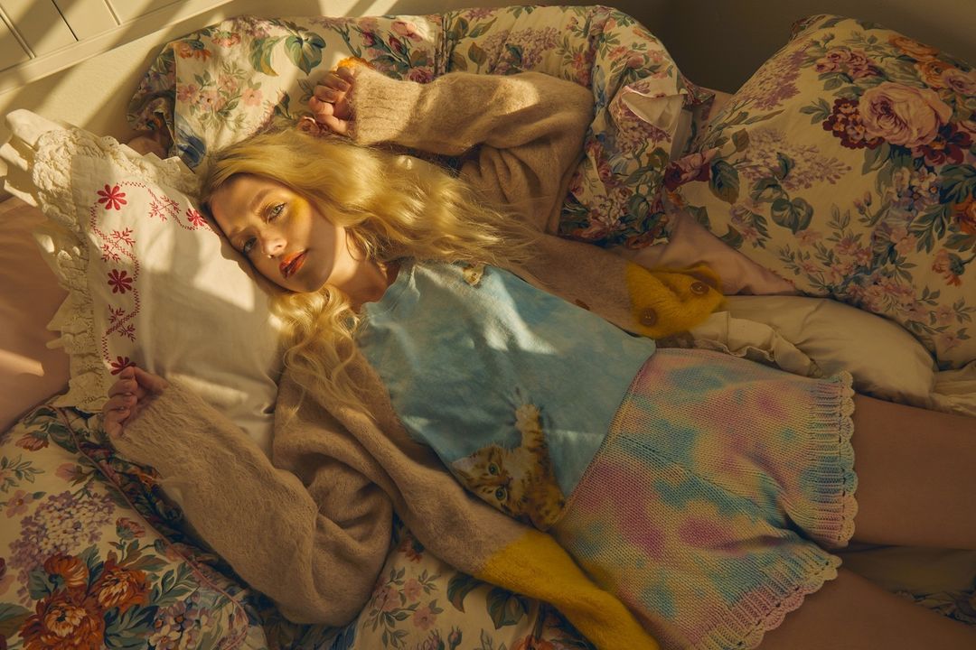 Blonde woman in light sweater lies on floral bedding in warm light. Soft and cozy scene with a relaxed and dreamy mood.