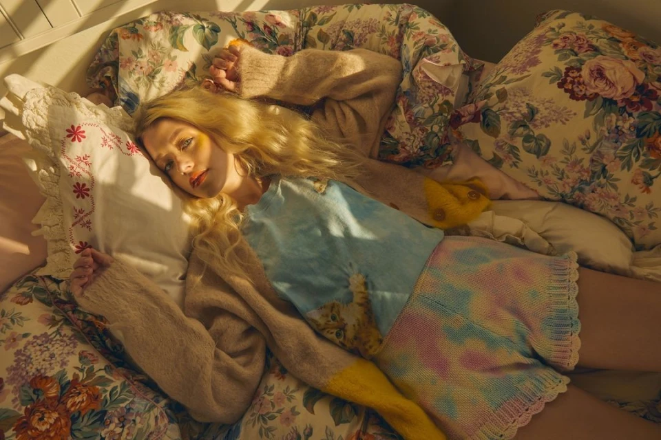 Blonde woman in light sweater lies on floral bedding in warm light. Soft and cozy scene with a relaxed and dreamy mood.