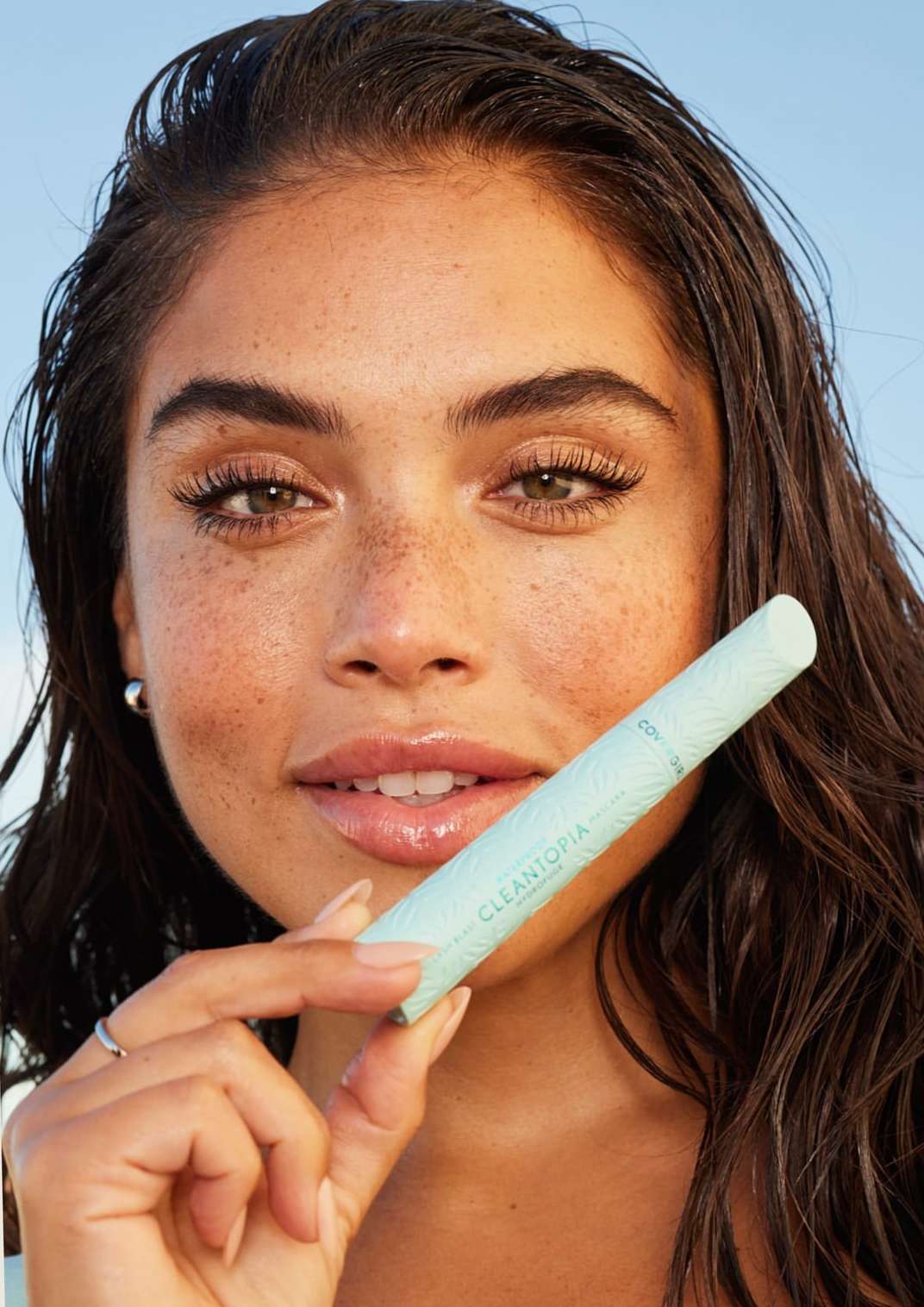 A young woman with freckles holds a pale green CLEANTOPIA mascara tube in front of a blurred outdoor backdrop, showcasing beauty and skincare.