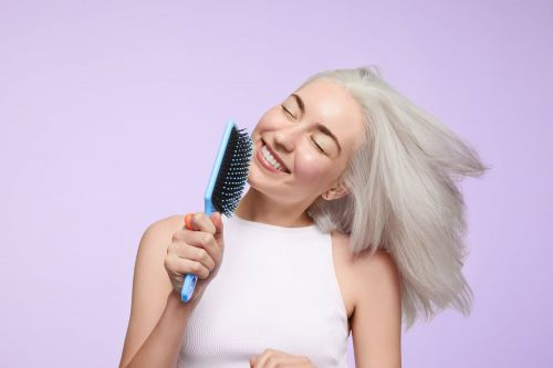 Smiling woman with gray hair holds a blue hairbrush and appears to be singing against a light purple backdrop in a medium shot.