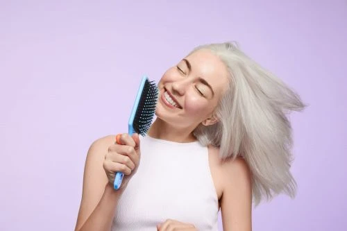 Smiling woman with gray hair holds a blue hairbrush and appears to be singing against a light purple backdrop in a medium shot.