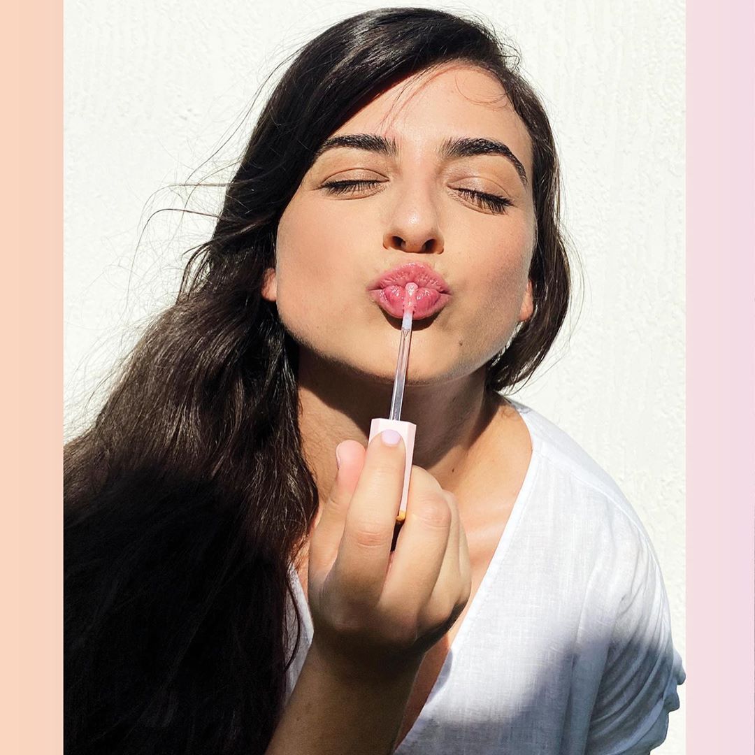 Brunette woman applies pink lip gloss with applicator wand in an eye-level shot on a white background, beauty advertisement.