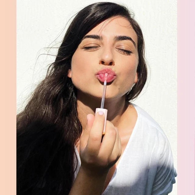 Brunette woman applies pink lip gloss with applicator wand in an eye-level shot on a white background, beauty advertisement.