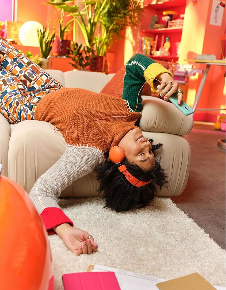 Woman relaxes on couch with orange headphones and phone in a colorful, plant-filled room. Bright, playful indoor setting.