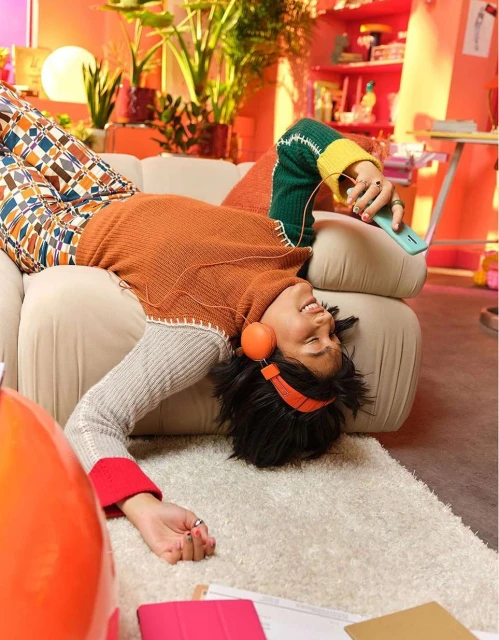 Woman relaxes on couch with orange headphones and phone in a colorful, plant-filled room. Bright, playful indoor setting.