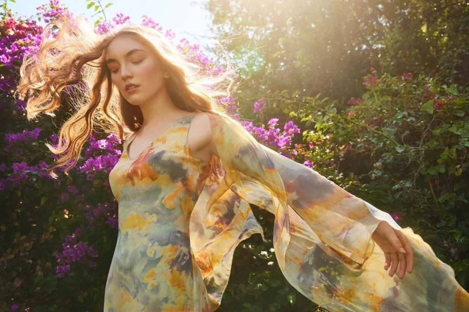 Young woman in a flowing dress poses in a lush garden with sun-drenched greenery and vibrant purple flowers during the day.