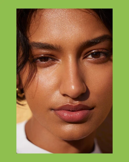 Close-up portrait of a smiling woman with brown hair, brown eyes, and smooth skin, wearing a white shirt against a green backdrop.