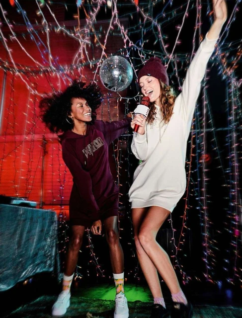 Two young women in a festive indoor setting with colorful string lights and a disco ball, one singing into a karaoke microphone.