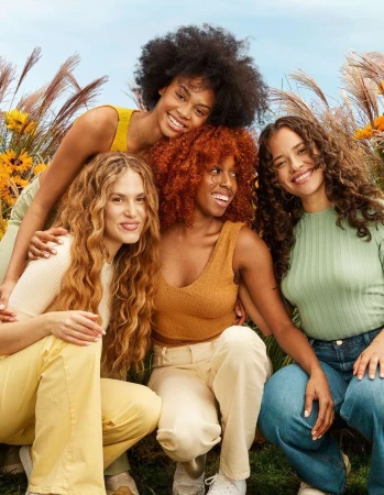 Group of four women smiling in outdoor setting with tall grasses and blue sky. Models with diverse hair styles pose together in a field.
