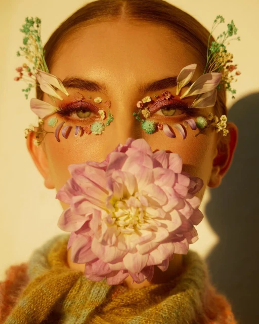 Woman with floral makeup, wearing a colorful sweater, and holding a large purple flower in front of her face, artistic portrait.
