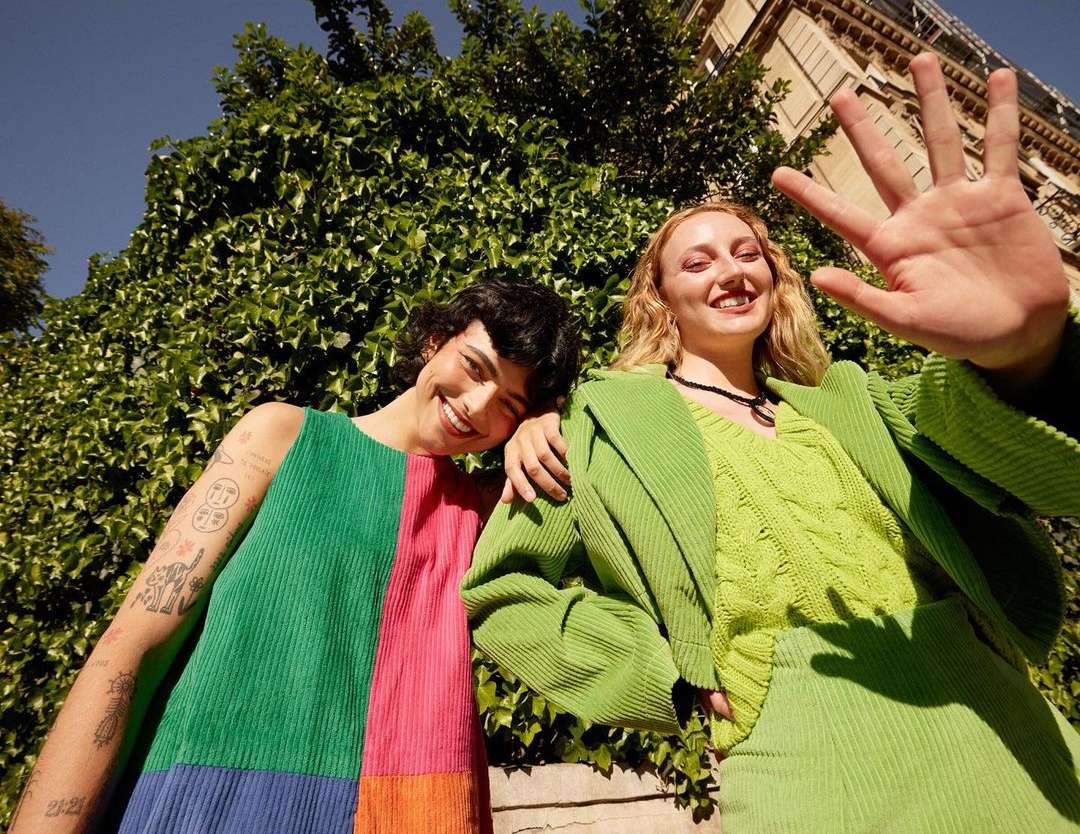 Two young adults, one in a colorful patchwork dress, the other in a green suit, smiling outdoors against a leafy wall and building.