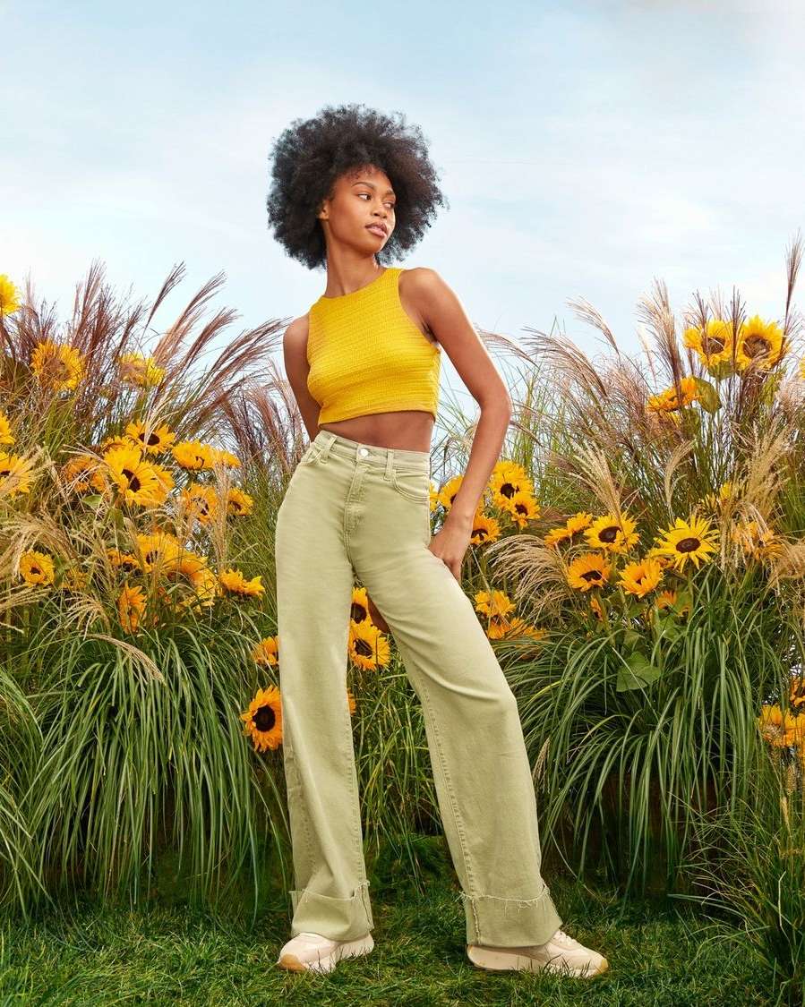 A young woman with Afro stands outside in a yellow knit top and sage-green, cuffed, wide-leg jeans surrounded by tall grasses and sunflowers.
