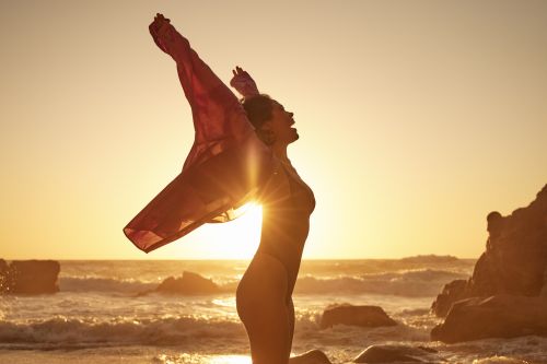 Woman stands with arms raised in backlight on a sandy beach at sunset holding sheer red fabric, ocean waves in the background.