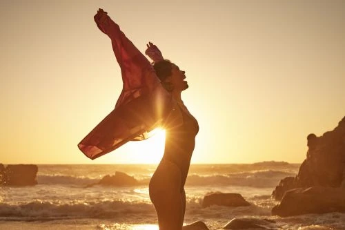 Woman stands with arms raised in backlight on a sandy beach at sunset holding sheer red fabric, ocean waves in the background.