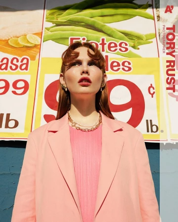A young woman poses outdoors in a pink outfit with a market sign background, under bright sunlight in a medium portrait shot.
