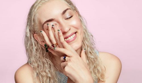 Smiling woman with light hair and colorful rings poses against a pink background in a close-up beauty shot.