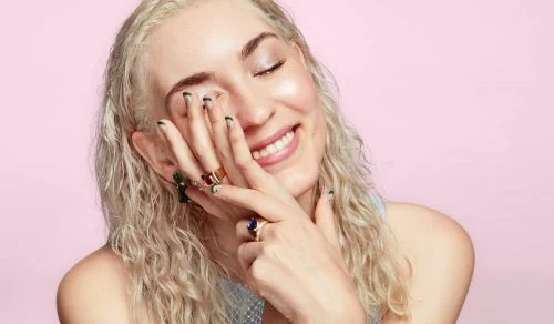 Smiling woman with light hair and colorful rings poses against a pink background in a close-up beauty shot.