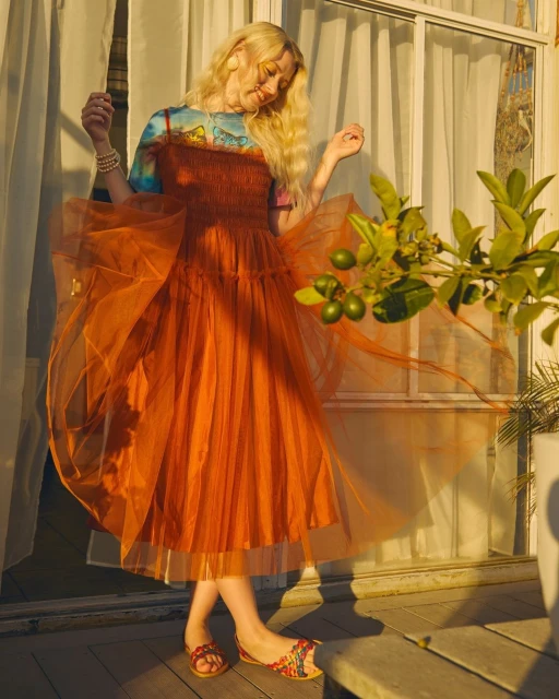 Young blonde woman in an orange tulle dress poses near a window with lime tree branches, bathed in warm light, creating a dreamy fashion image.
