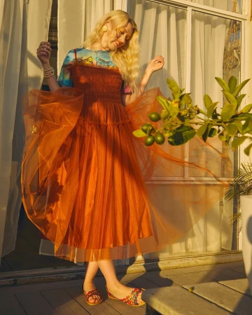 Young blonde woman in an orange tulle dress poses near a window with lime tree branches, bathed in warm light, creating a dreamy fashion image.