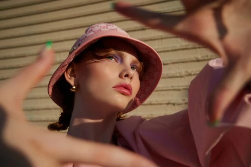 Close-up of a fair-skinned woman wearing a pink bucket hat and shirt, framing her face with her hands, against a neutral background.