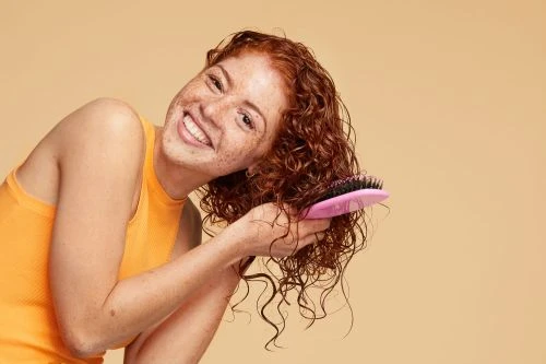 Smiling woman with curly red hair brushes it to the side with a pink hairbrush, wearing a tank top on a beige background.