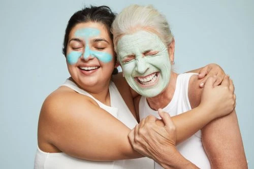 Two women with facial masks smile and embrace against a light blue background, showcasing skincare and joyful connection.
