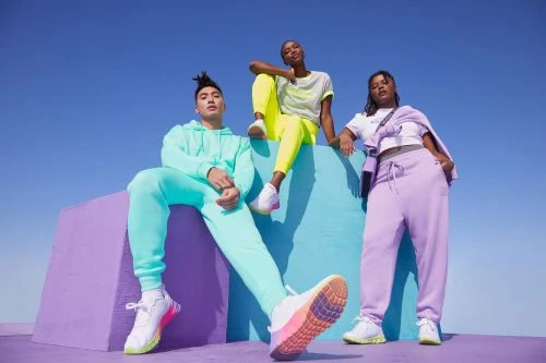 Three adult models showcase pastel-colored athletic wear and sneakers on a colorful geometric platform against a bright blue sky.