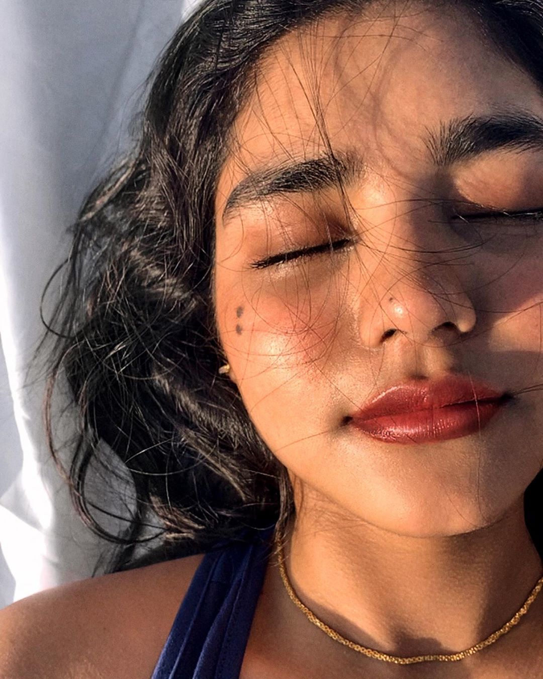 Close-up of a young adult woman with closed eyes, dark hair, mole on her face, gold necklace, and a blue top, basking in natural light.