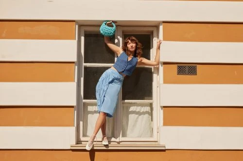 Young woman in blue outfit poses dynamically in a window frame with an orange and white striped wall backdrop, holding a blue purse.