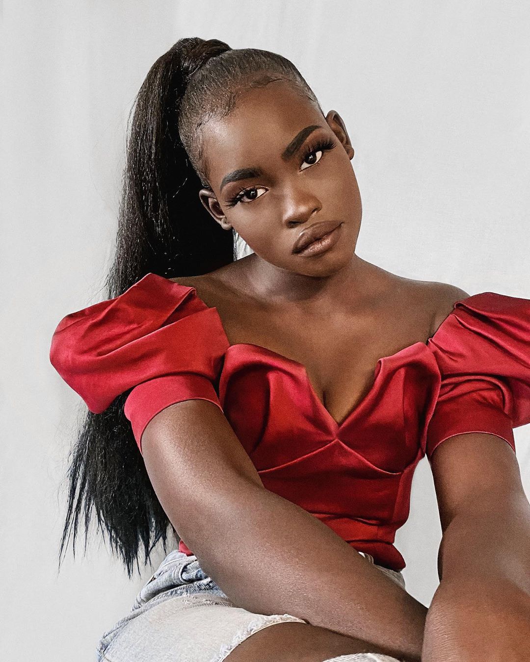 Portrait of a young, dark-skinned woman in a red puff-sleeve top and denim shorts against a white background, with a high ponytail.