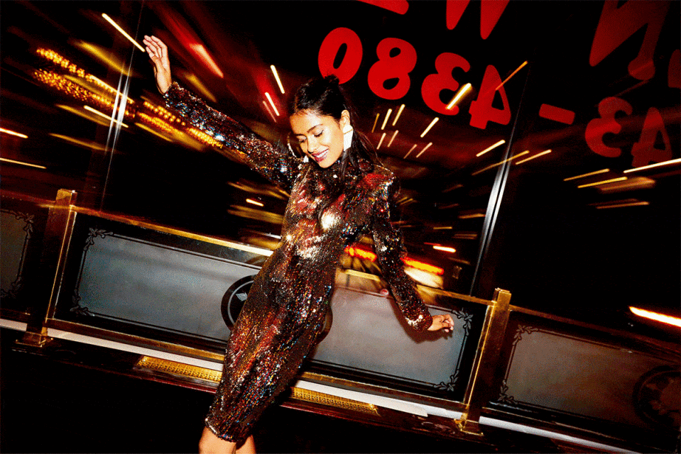 Woman in shimmering bronze dress poses indoors with dark background and blurred lights, smiling, expressing energy and elegance in a stylish setting.