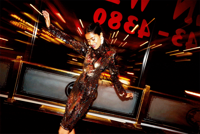 Woman in shimmering bronze dress poses indoors with dark background and blurred lights, smiling, expressing energy and elegance in a stylish setting.