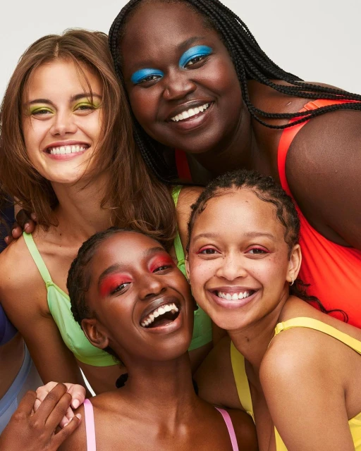 Close-up of four smiling diverse women with colorful makeup, wearing brightly colored tops against a white backdrop.