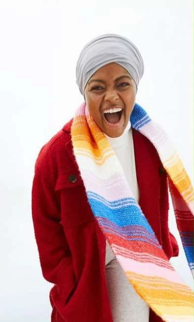 A woman with a headscarf smiles widely, wearing a red jacket and a colorful striped scarf against a bright background.