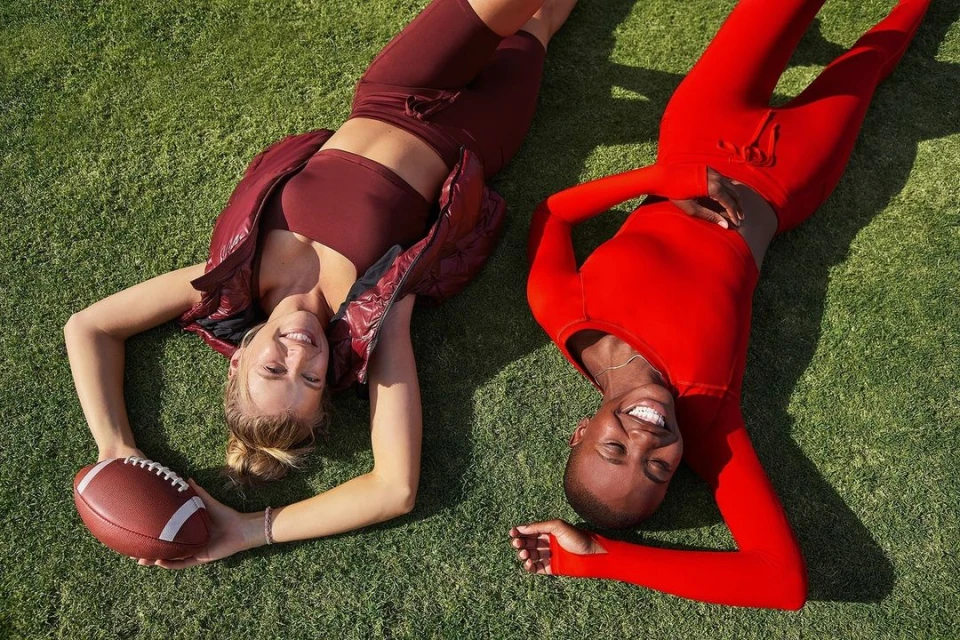 Two women lie on a grass field in sportswear, smiling and holding a football, illuminated by natural sunlight, enjoying leisure outdoors.