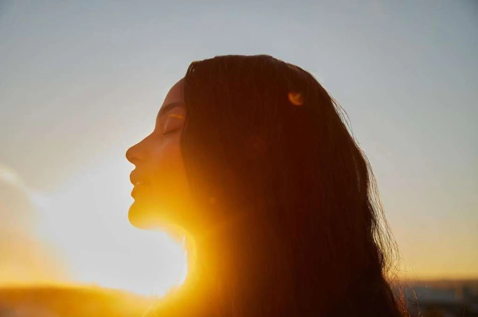 Profile portrait of an adult female with long dark hair looking to the left with eyes closed and sun shining behind her in an outdoor setting.
