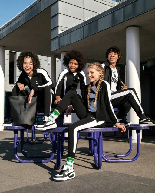 Four young people in Puma tracksuits sit on a purple table outdoors. The shot captures a lifestyle moment with an urban background.