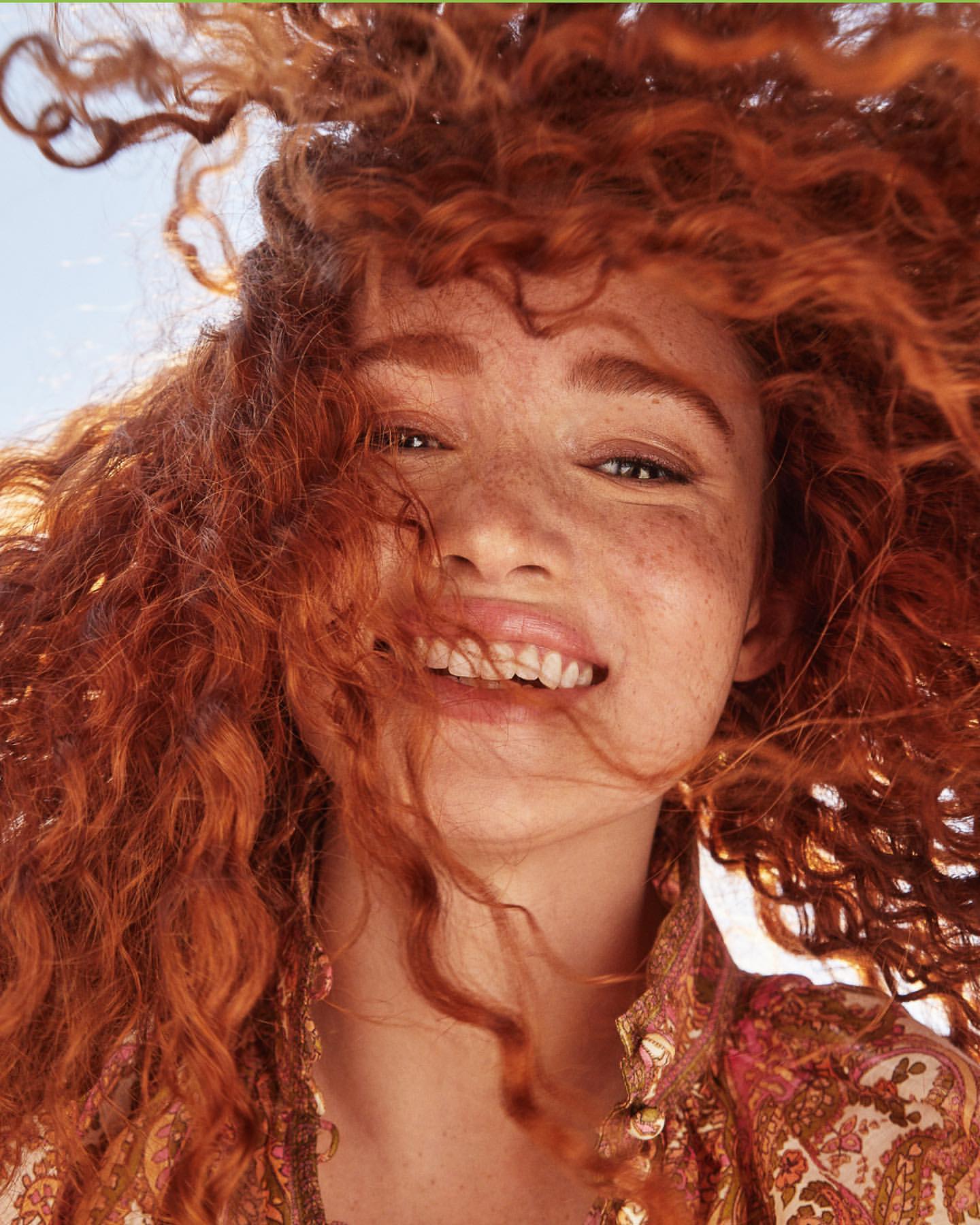 Close-up of a smiling red-haired woman with freckles looking up toward the camera in a brightly lit, sunny outdoor setting.