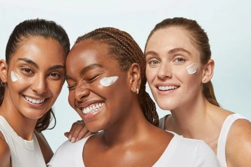Three diverse women smiling with cream on their faces, skincare concept. Close-up shot of models against light-blue studio backdrop.
