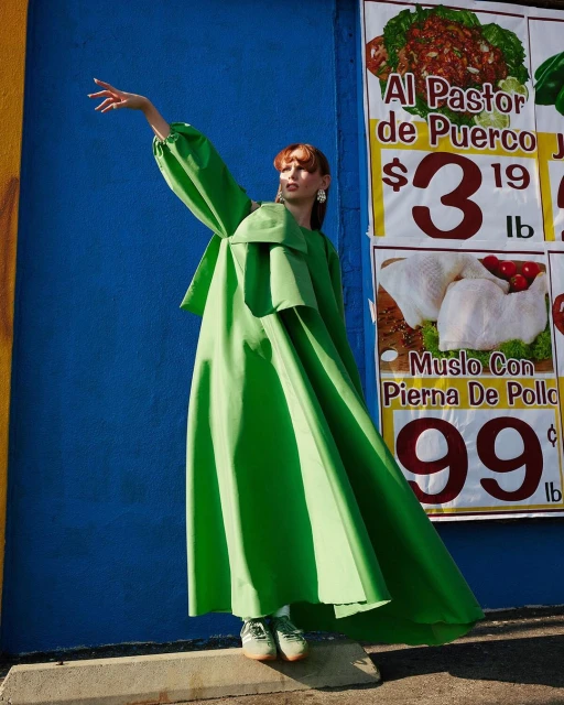 A woman in a flowing green dress poses in front of a blue wall with colorful food advertisements, presenting a striking and fashionable image.