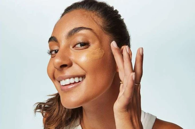 Close-up portrait of a woman applying foundation to her cheek against a clean white background, photographed by Tory Rust.
