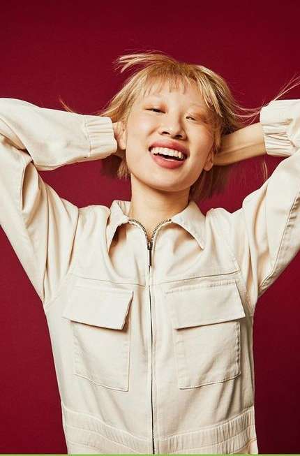 Smiling woman with short blonde hair, wearing a beige jumpsuit, poses against a red background with hands behind her head.