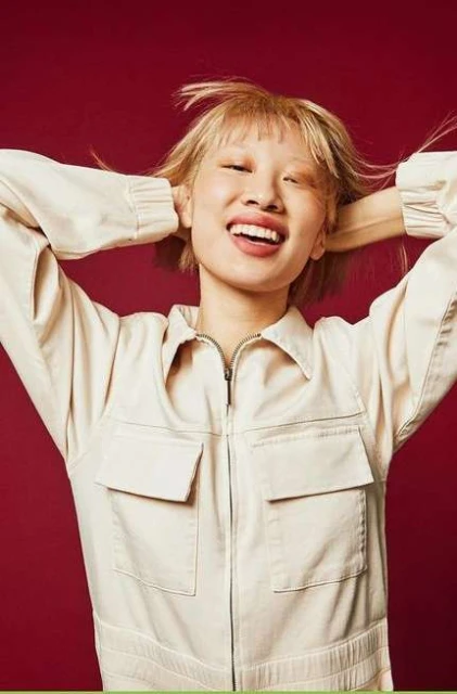 Smiling woman with short blonde hair, wearing a beige jumpsuit, poses against a red background with hands behind her head.