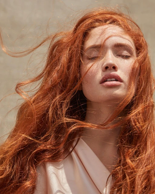 Close-up of a red-haired woman with her eyes closed, wearing a light pink shirt, against a neutral background with hair blowing.