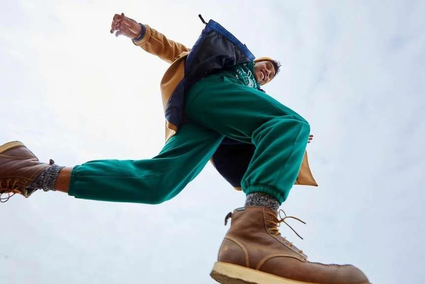 A man with a tan jacket, green sweatpants, and brown boots appears to jump in the air, captured from a low angle against a bright sky.