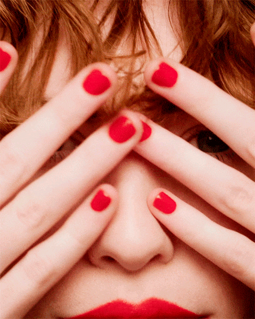 Close-up portrait of a young adult woman with red hair and bright red manicure, covering her face with her hands.